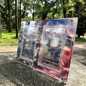 K&ouml;nnte beinhalten: Eine transparente Acryl-Vitrine mit zwei bewerteten Pok&eacute;mon-Sammelkarten. Die Karten befinden sich in Schutzh&uuml;llen mit Etiketten. Der Hintergrund hat ein farbenfrohes, abstraktes Design. Das Geh&auml;use ist mit silberner Hardware befestigt.