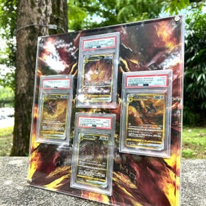 Puede incluir: Una pantalla acr&iacute;lica transparente con cinco cartas coleccionables de Pok&eacute;mon en estuches protectores. Las cartas tienen ilustraciones doradas y marrones. El fondo tiene un dise&ntilde;o ardiente con tonos rojos, naranjas y amarillos. Las cartas est&aacute;n etiquetadas.