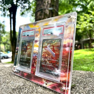 May include: A clear acrylic display case holding two Pokemon trading cards. The cards feature a red and white Latias character with Japanese text. The case is set against a colorful sunset background.