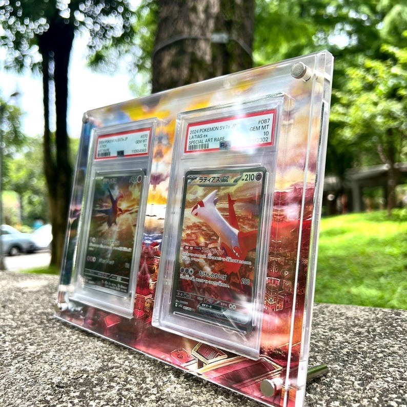 May include: A clear acrylic display case holding two Pokemon trading cards. The cards feature a red and white Latias character with Japanese text. The case is set against a colorful sunset background.