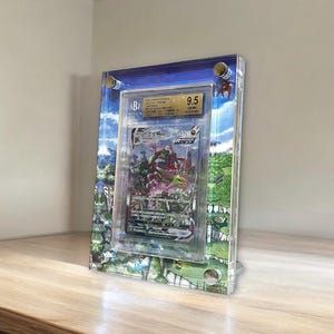 May include: A graded trading card in a clear acrylic frame. The frame has a landscape design with blue sky and green fields. The card is graded 9.5 and features a colourful illustration.