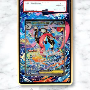 Könnte beinhalten: Eine PSA-zertifizierte Mega Greninja EX Sammelkarte in einer transparenten Kunststoffhülle mit goldenem Rand. Die Karte zeigt eine farbenfrohe Illustration der blau-schwarzen Figur mit japanischem Text vor einem wirbelnden kosmischen Hintergrund.