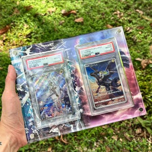 K&ouml;nnte beinhalten: Zwei Pok&eacute;mon-Sammelkarten in Schutzh&uuml;llen. Eine Karte zeigt Reshiram, die andere Zekrom. Beide Karten sind von PSA als Gem Mint 10 bewertet. Der Hintergrund ist ein farbenfrohes, abstraktes Design.