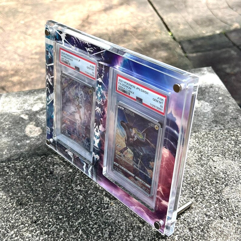 K&ouml;nnte beinhalten: Transparenter Acryl-Display-Rahmen mit zwei bewerteten Pok&eacute;mon-Sammelkarten. Der Rahmen hat einen Metallst&auml;nder und ist mit kleinen silbernen Schrauben befestigt. Die Karten sind mit ihren jeweiligen Bewertungsaufklebern sichtbar.