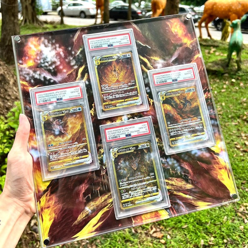 Puede incluir: Colecci&oacute;n de cuatro cartas coleccionables de Pok&eacute;mon, encapsuladas en estuches protectores. Las cartas presentan ilustraciones doradas y negras con texto japon&eacute;s, sobre un fondo de fuego. Se exhiben en una placa acr&iacute;lica transparente.