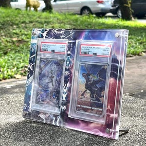 K&ouml;nnte beinhalten: Ein klarer Acryl-Displayrahmen mit zwei Pok&eacute;mon-Sammelkarten. Die Karten befinden sich in Schutzh&uuml;llen, mit sichtbarem Text wie "N'S RESHIRAM" und "ZEKROM VMAX". Der Rahmen hat einen farbenfrohen, abstrakten Hintergrund.