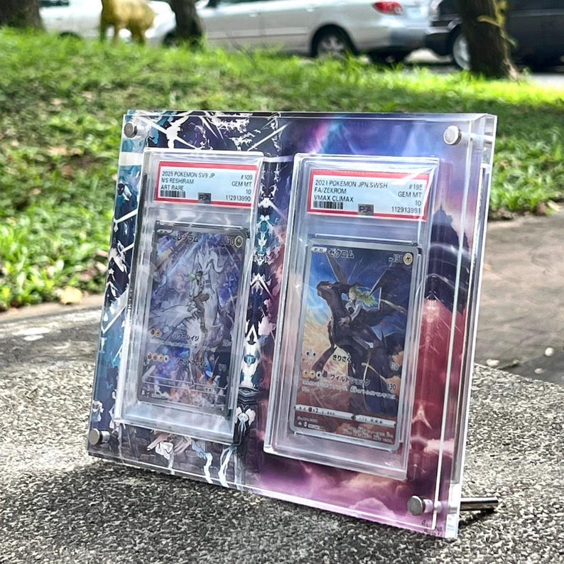 K&ouml;nnte beinhalten: Ein klarer Acryl-Displayrahmen mit zwei Pok&eacute;mon-Sammelkarten. Die Karten befinden sich in Schutzh&uuml;llen, mit sichtbarem Text wie "N'S RESHIRAM" und "ZEKROM VMAX". Der Rahmen hat einen farbenfrohen, abstrakten Hintergrund.