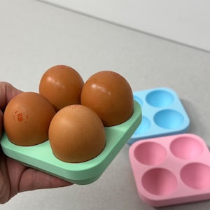 Könnte beinhalten: Ein hellgrüner Eierhalter mit vier braunen Eiern. Zwei weitere Eierhalter sind sichtbar, einer hellblau und einer rosa. Die Eier haben einen rotbraunen Stempel.