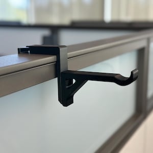 Op de afbeelding: Een zwarte metalen haak bevestigd aan een grijs bureau of scheidingswand. De haak is ontworpen om items op te hangen en wordt met een klem gemonteerd. De haak is gemaakt van een stevig materiaal en is geschikt voor kantoor of thuis.