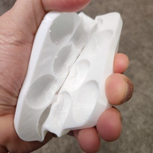 May include: White plastic mold with multiple oval-shaped cavities, split in half and held open by a hand.