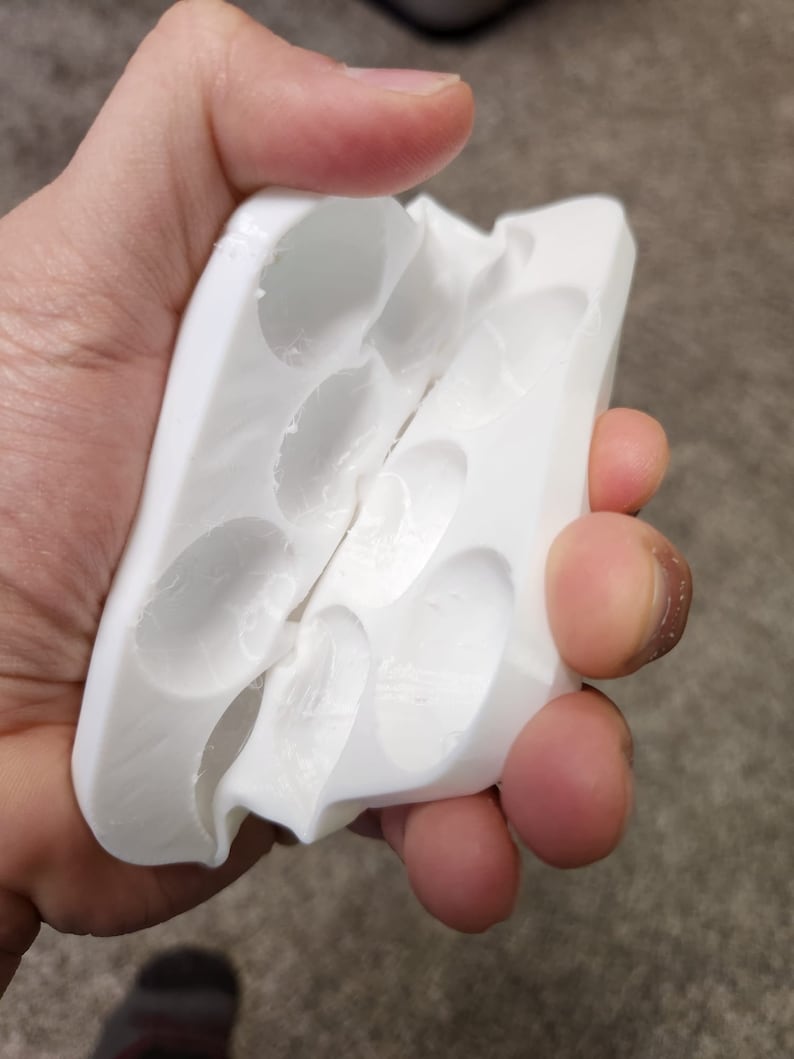 May include: White plastic mold with multiple oval-shaped cavities, split in half and held open by a hand.