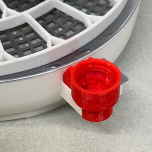 May include: Close-up of a white and gray incubator with a red water fill port. The incubator has a circular design with multiple compartments and a clear top. The red port is on the side of the incubator.