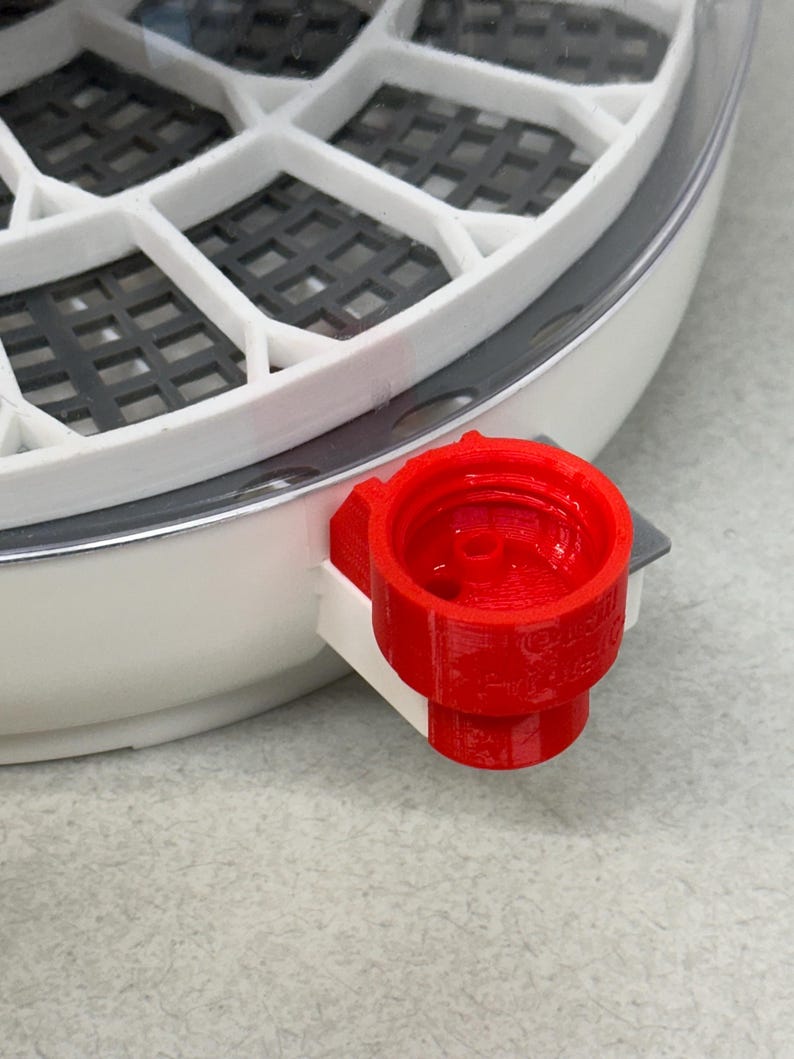 May include: Close-up of a white and gray incubator with a red water fill port. The incubator has a circular design with multiple compartments and a clear top. The red port is on the side of the incubator.