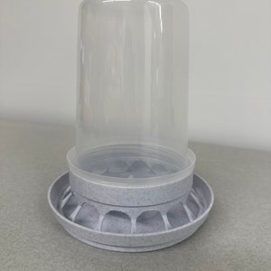 Peut inclure: Un distributeur d'aliments pour oiseaux en plastique blanc avec un récipient en plastique transparent sur le dessus. Le distributeur a une base circulaire avec une surface texturée et une série de trous autour du bord.