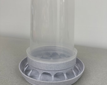 Puede incluir: Un comedero para pájaros de plástico blanco con un recipiente de plástico transparente en la parte superior. El comedero tiene una base circular con una superficie texturizada y una serie de agujeros alrededor del borde.