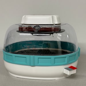 May include: A white and teal electric food dehydrator with a clear plastic dome lid. The dehydrator has a red button on the side and the text "OurGreenHome" on the front.
