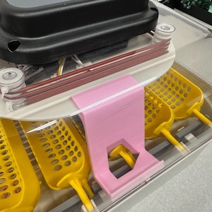 Op de afbeelding: Een witte plastic incubator met een doorzichtig deksel en gele eierrekjes. Een roze plastic beugel is aan de zijkant van de incubator bevestigd. De incubator heeft een digitaal display met de tekst "MATICOO" aan de voorkant.