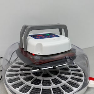 May include: A white and gray egg incubator with a clear, domed lid. The incubator has a digital display and a gray handle. The base is white with a black grid pattern. The incubator is designed for hatching eggs.