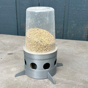 Op de afbeelding: Een grijze plastic vogelvoeder met een doorzichtige plastic container gevuld met vogelzaad. De voeder heeft drie poten en een ronde basis met gaten voor de vogels om toegang te krijgen tot het zaad.