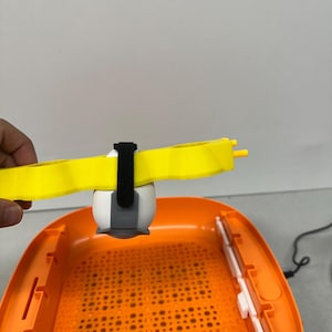 May include: An orange and white egg incubator with a yellow support arm and a white egg. The incubator has a perforated base and white interior components. A black power cord is visible.