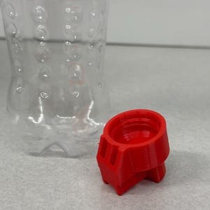 May include: A clear plastic bottle with a textured surface and a bright red 3D-printed cap. The cap is cylindrical with a geometric design. The bottle is empty and has a standard shape.
