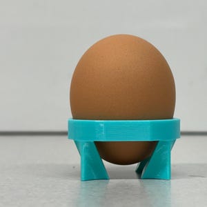 May include: A brown egg sits in a turquoise egg holder. The egg holder is a small, three-legged stand with a circular top designed to cradle the egg. The egg is a light brown color and appears to be a standard size.