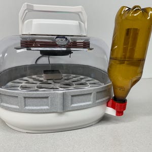 May include: A white and gray incubator with a clear dome and a brown bottle attached. The incubator has a handle and a circular base with a grid pattern. The bottle is brown and connected to a red and white valve.