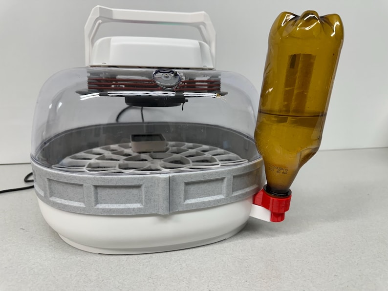 May include: A white and gray incubator with a clear dome and a brown bottle attached. The incubator has a handle and a circular base with a grid pattern. The bottle is brown and connected to a red and white valve.