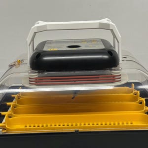 May include: An egg incubator with a clear, curved lid, a black base, and yellow egg trays. It features a black control panel with buttons and a white handle. The incubator is designed for hatching eggs.