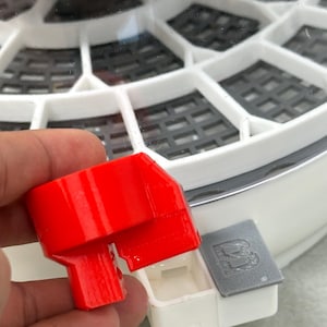 May include: A bright red plastic component, possibly a part of a larger device, held in a hand. The component has a rectangular shape with a protruding section. A small, gray rectangular piece with a logo is next to it.