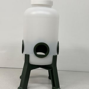 May include: A white plastic bird feeder with a screw-on lid and three circular openings. The feeder is supported by a dark green, three-legged stand. The feeder is designed to hold birdseed and attract birds to a garden or outdoor space.