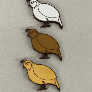 Op de afbeelding: Drie houten uitsnijdingen van kwartels in verschillende kleuren: wit, bruin en goud. De vogels kijken naar links en hebben een eenvoudig, gestileerd ontwerp.