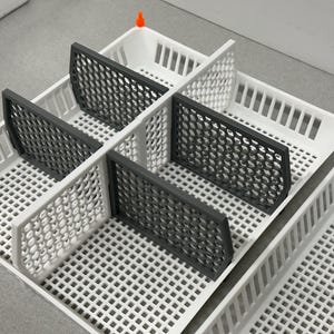 May include: A white plastic storage container with multiple grey dividers. The container has a grid pattern and is designed to hold small items.