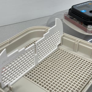 Op de afbeelding: Een witte kunststof eierenbroeder met een witte gerasterde lade en een witte kunststof scheidingswand. De broeder heeft een transparante kunststof koepel bovenop met een digitaal bedieningspaneel met het label "MATICOOOPY".