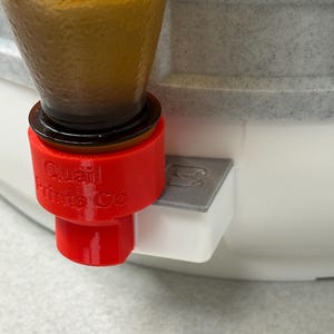 May include: A red 3D-printed adapter with the text "Quail Prints Co" attached to a clear glass bottle. The adapter is connected to a white and gray container. The bottle contains a brown liquid.