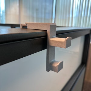 May include: A white plastic hook mounted on a black cubicle wall. The hook has two prongs for hanging items.