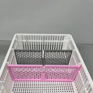 May include: A white plastic storage basket with two grey and two pink dividers. The dividers have a honeycomb pattern.