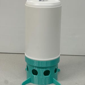 Op de afbeelding: Een witte en teal cilindrische vogelvoeder. De bovenkant en het hoofdgedeelte zijn wit, terwijl de basis en de poten teal zijn. De basis heeft verschillende ronde openingen. De feeder heeft een modern, minimalistisch ontwerp.