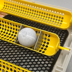 May include: A close-up of an incubator with yellow egg trays and a speckled egg. The trays have a grid pattern of holes. The egg is light gray with darker speckles. The incubator has a black mesh base.