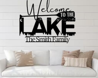 Puede incluir: Arte de pared de metal negro con las palabras "Welcome to the Lake" y "The Smith Family". La palabra "Lake" está estilizada con una paleta y estrellas. El arte está montado en una pared blanca sobre un sofá blanco con cojines.