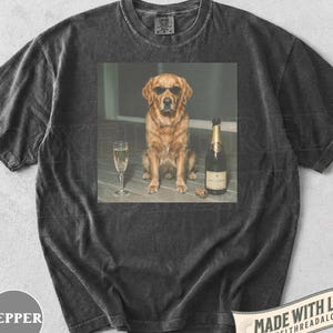 May include: A charcoal gray short-sleeve t-shirt featuring a graphic print of a golden retriever wearing black sunglasses, sitting next to a champagne flute and a bottle of champagne with a cork lying on the surface near the bottle base.