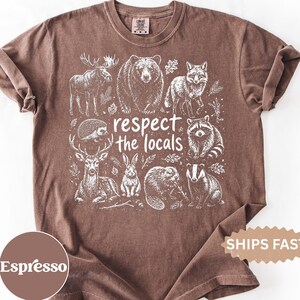 Könnte beinhalten: Ein braunes Kurzarm-T-Shirt mit einem weißen Grafikdruck verschiedener Waldtiere, darunter ein Elch, ein Bär, ein Fuchs, ein Igel, ein Hirsch, ein Hase, ein Biber und ein Dachs, mit dem mittig platzierten Text "respect the locals".