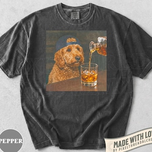 K&ouml;nnte beinhalten: Dunkelgraues T-Shirt mit einem quadratischen Grafikdesign. Die Grafik zeigt einen Golden Doodle mit blauer Kappe, der ein Glas Whiskey mit Eis betrachtet. Whiskey wird in das Glas gegossen. Das T-Shirt hat einen Vintage-Look.