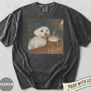 May include: Dark gray t-shirt featuring a graphic of a white dog wearing a blue baseball cap, sitting at a table while beer is poured into a mug. The shirt has a vintage, washed look. The text "MADE WITH LOVE" is visible.