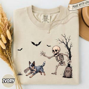 May include: A beige t-shirt featuring a graphic print of a skeleton running from a dog that is holding a bone in its mouth. The design includes a tombstone labeled RIP, a bare tree, and three flying bats. The shirt has a crew neckline and short sleeves.