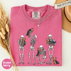 May include: A pink crewneck t-shirt featuring a graphic print of four human skeletons interacting with small brown and black Yorkshire Terrier dogs. The shirt has a Comfort Colors brand label on the inside neck and a star-shaped silver necklace.