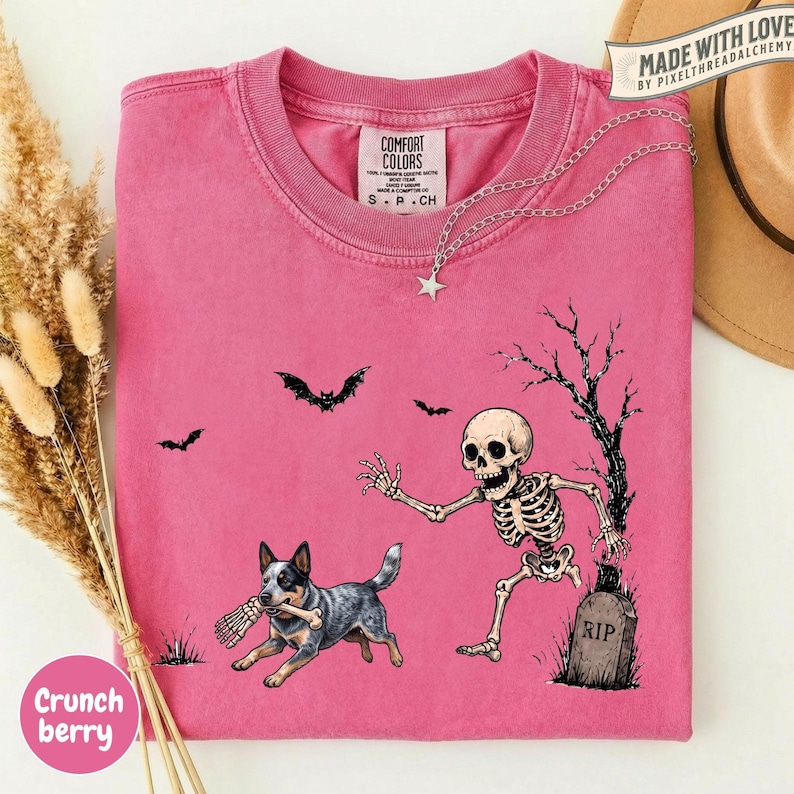 May include: A pink short-sleeve t-shirt featuring a graphic of a skeleton running from a tombstone labeled RIP, a dog holding a bone, and two bats. The shirt has a crew neckline and a Comfort Colors brand label printed on the inside collar.