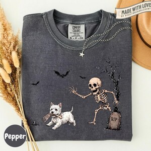 May include: A charcoal gray crew neck t-shirt featuring a graphic print of a skeleton reaching toward a white West Highland White Terrier dog holding a bone in its mouth. The design includes a tombstone labeled RIP, a bare tree, and bats.