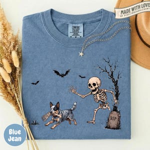 May include: A blue t-shirt featuring a graphic of a skeleton running toward a tombstone marked RIP, while a dog carries a bone in its mouth. Two bats fly above the scene, and a bare tree stands next to the grave. The shirt has a crew neckline.