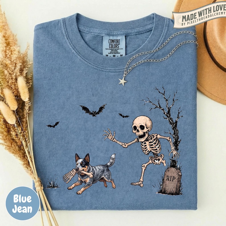 May include: A blue t-shirt featuring a graphic of a skeleton running toward a tombstone marked RIP, while a dog carries a bone in its mouth. Two bats fly above the scene, and a bare tree stands next to the grave. The shirt has a crew neckline.